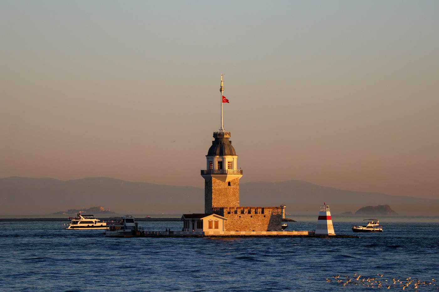 Maiden's Tower on its islet in the Bosphorus at golden hour, with small boats passing and the hills of the Asian shore behind